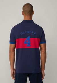 Camisa polo navy con una franja roja, que presenta "HACKETT" y el número "4" en azul en la parte posterior. Diseño de manga corta y con cuello.