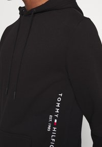 Sudadera negra con capucha y cordones, con texto vertical blanco "Tommy Hilfiger Est. 1985" y un pequeño logo de bandera en rojo, blanco y azul en el costado.