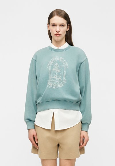 Femme portant un sweat-shirt vert sarcelle avec l'impression "Fontaines de Paris", une chemise blanche à col en dessous, et un short beige, debout contre un fond uni.