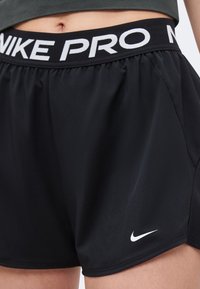 Κοντινό ενός ατόμου που φοράει μαύρα σορτς Nike Pro με λευκό λογότυπο και ελαστική μέση πάνω από τον αφαλό.
