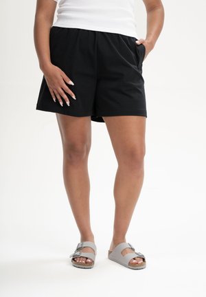 Personne portant un short ample noir, un haut sans manches blanc et des sandales à double lanière gris clair, debout devant un fond blanc.