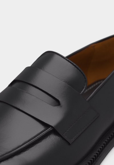 Κοντινό πλάνο μαύρου δερμάτινου μοκασίνι με λεία επιφάνεια και λεπτομέρεια ρουχισμού penny strap, δείχνοντας το πάνω μέρος του παπουτσιού και μέρος της εσωτερικής επένδυσης.
