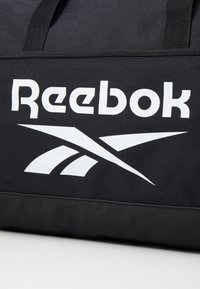 Μαύρη τσάντα ντουφλέ από ύφασμα με έντονο λευκό λογότυπο "Reebok" και δυναμικό στοιχείο γραφιστικού σχεδιασμού. Με φερμουάρ στο πάνω μέρος.