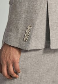 Hellgraue Leinenjacke mit vier hellbeigen Knöpfen an den Ärmeln, strukturiertem Stoff und taillierter Passform. Die Hand ruht auf dem Hosenbein.