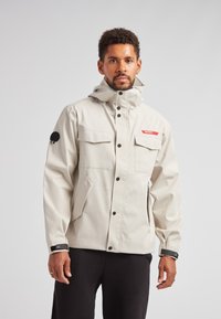 Lys beige vandtæt jakke med hætte, to frontlommer og et rødt logo. Bemærkelsesværdig sort lap på ærmet, justerbare manchetter med Velcro.