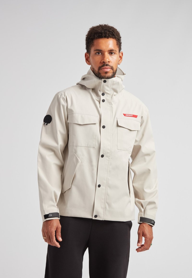 Lys beige vandtæt jakke med hætte, to frontlommer og et rødt logo. Bemærkelsesværdig sort lap på ærmet, justerbare manchetter med Velcro.