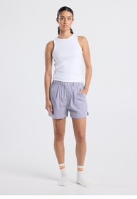 Valkoinen hihaton toppi yhdistetty sinivalkoiseen gingham-kuvioisiin shortseihin, joissa on nappikiinnitys ja sivutaskut, käytetty raidallisten sukkien kanssa.