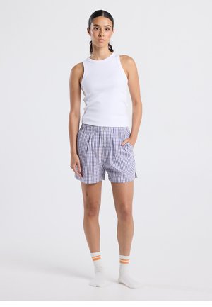 Weißes ärmelloses Tanktop kombiniert mit Shorts im blau-weißen Vichy-Karo-Muster, ausgestattet mit Knopfverschluss und Seitentaschen, getragen mit gestreiften Socken.