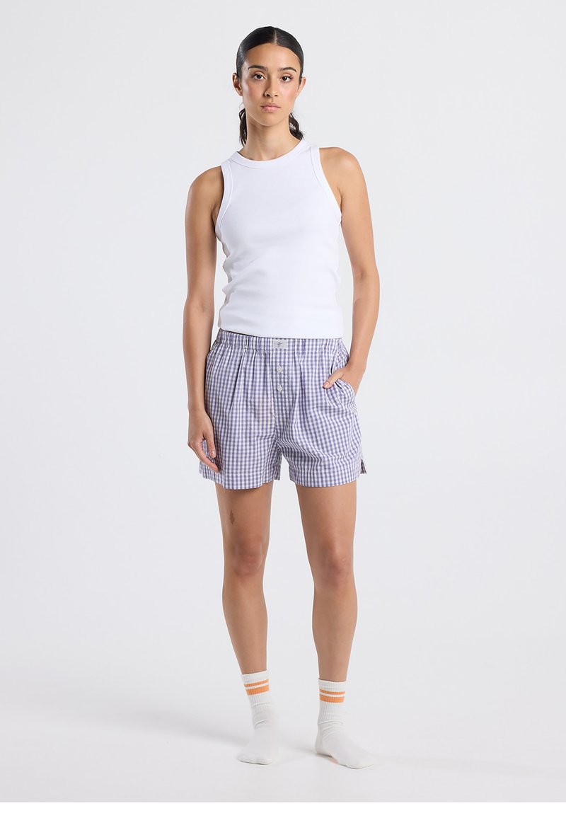 Valkoinen hihaton toppi yhdistetty sinivalkoiseen gingham-kuvioisiin shortseihin, joissa on nappikiinnitys ja sivutaskut, käytetty raidallisten sukkien kanssa.