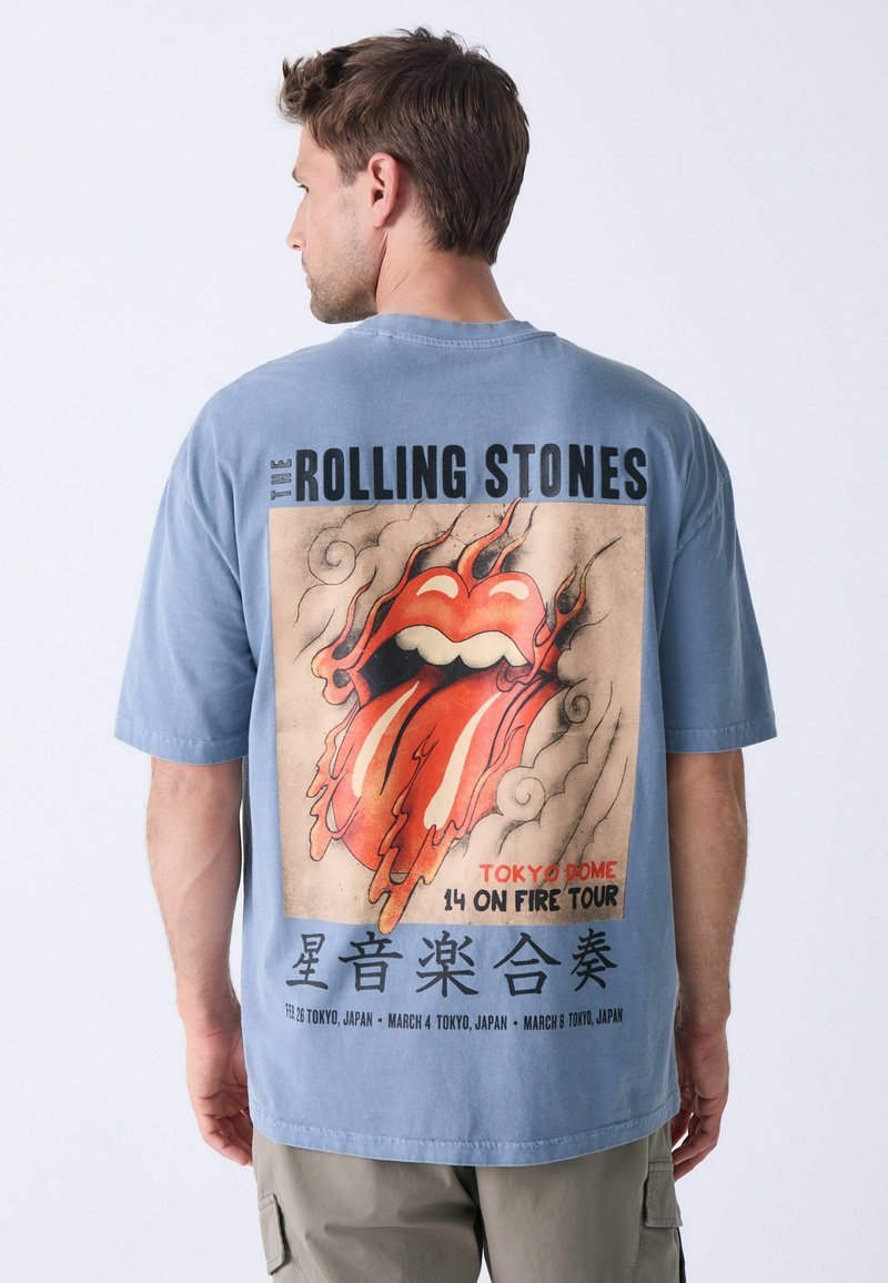 Svijetloplavi pamučni majica s velikim grafičkim prikazom logotipa Rolling Stonesa, s plamenovima i natpisom koji detaljno opisuje koncertnu turneju u Japanu.