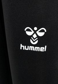 Hvid stiliseret humlebi-logo og teksten "hummel" trykt på sort stof, sandsynligvis sportstøj.