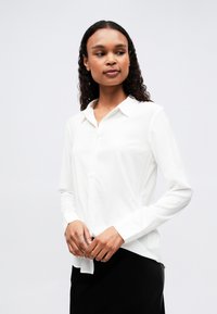 Chemise blanche à col, à manches longues et en tissu lisse ; assortie à une jupe noire, mettant en avant un design classique et minimal.