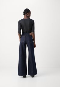 Vide benedørk jeans med høy liv, i mørk denim, med store baklommer og en brun lærpatch. En svart langermet topp er brukt.