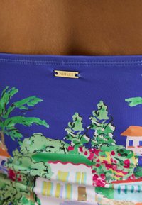Tissu bleu avec imprimé coloré d’arbres tropicaux et de maisons, avec une petite étiquette métallique portant le nom de la marque « Joules » cousue près du bord supérieur.