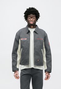 Šedá bavlnená bunda s golierom, s červenými a modrými vyšívanými nášivkami na hrudi a rukáve a krémovými bočnými panelmi.