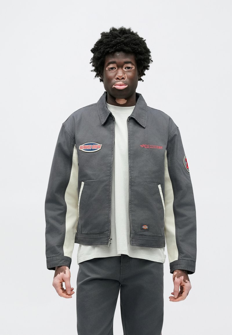 Gray cotton jacket with a collar, featuring red and blue embroidered patches on the chest and sleeve, and cream-colored side panels.