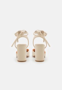 Beige høyhælte sandaler med firkantet tå, blokkhel og knyttede ankelstropper. Glatt tekstur med en brun såle-akse.