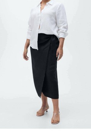 Mujer con camisa blanca abotonada, falda negra cruzada y tacones abiertos color nude, de pie frente a un fondo blanco liso.