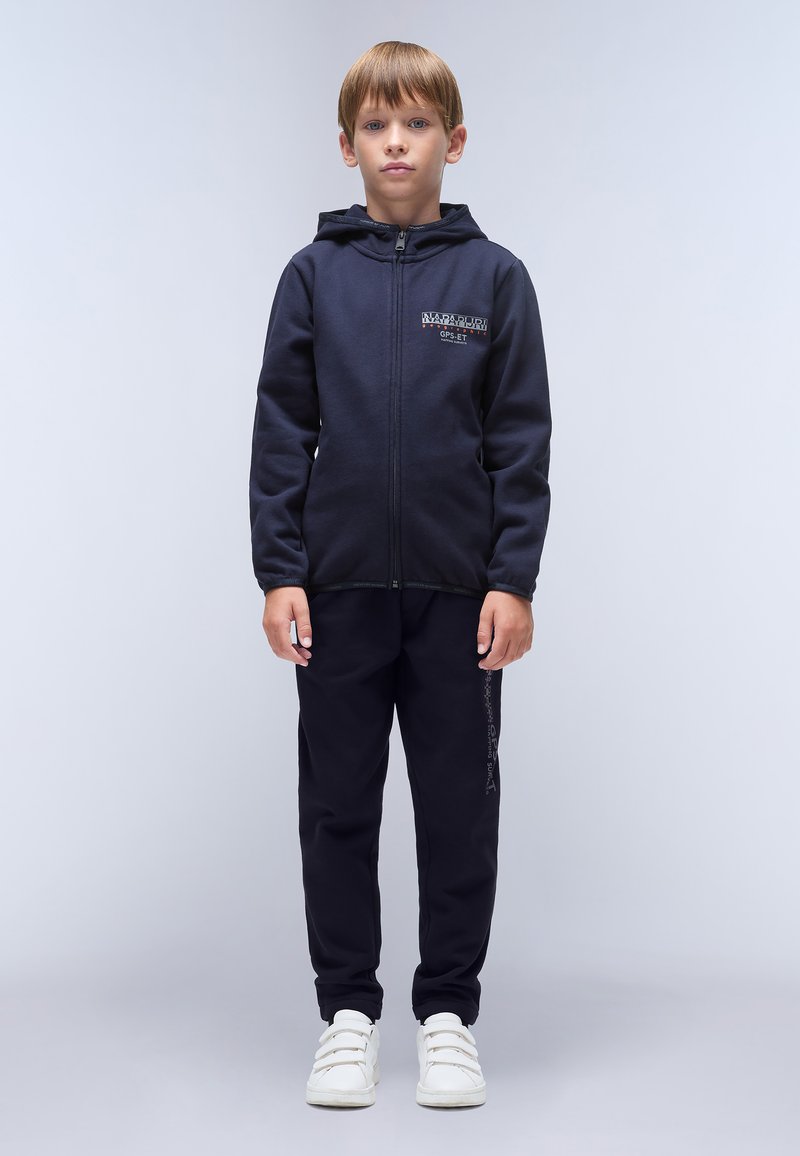 Marineblå zip-hoodie med logo-detalje, parret med matchende joggers og hvide sneakers. Har lange ærmer og en frontlynlås.