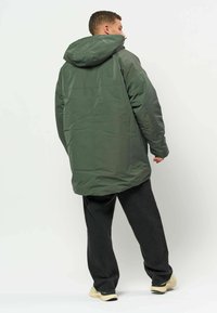 Homme debout de dos, portant une veste à capuche vert foncé, un pantalon noir et des chaussures claires sur fond uni.