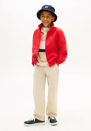 Ragazzo sorridente che indossa una giacca rossa, polo beige, pantaloni, cappello a tesa larga blu navy e sneakers blu navy con dettagli bianchi, in piedi con le mani nelle tasche.