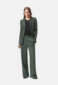 Traje verde hecho a medida con blazer de corte slim y pantalones de pierna ancha, combinado con una camiseta negra ajustada con letras blancas. Textura de tela suave.