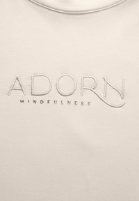 Helles beiges Gewebe mit gesticktem Text "ADORN" über dem kleineren "MINDFULNESS" in Grau. Glatte Textur und Rundhalsausschnitt.