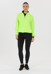 Neongrön lättviktsjacka med dragkedja framtill och svart krage, parat med svarta leggings med prickiga detaljer och svarta sneakers.