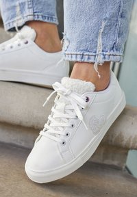 Sneaker bianchi con polsini alla caviglia in tessuto peloso, punta tonda e chiusura con lacci. Presentano un delicato accento a forma di cuore sul lato. Cuffs in denim visibili.