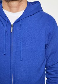 Blaues Zip-Hoodie aus strukturiertem Strick, mit zwei Zugbändern und einem Reißverschluss. Trägt ein weißes T-Shirt am Hals darunter.