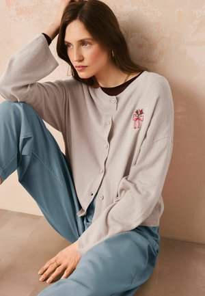 Frau mit langen braunen Haaren sitzt an einer strukturierten pfirsichfarbenen Wand und trägt eine hellgraue Strickjacke mit Blumenstickerei und lockere blaue Hosen.