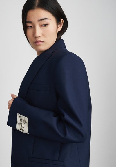 Marineblauer Blazer aus strukturiertem Stoff, mit einem einreihigen Design, Reverskragen und einem Etikett am Ärmel.