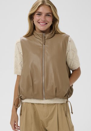 Mosolygó nő bézs cipzáras műbőr mellényben, krémszínű rövid ujjú kötött pulóver fölött, és világosbarna magas derekú pliszírozott nadrágban.