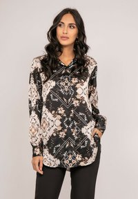 Blouse à manches longues noire et crème, ornée de motifs floraux et géométriques, avec un devant à boutons et un col montant. Texture satin lisse.