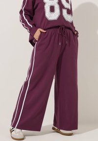 Bordeauxfarbene Sweatpants mit entspannter Passform, ausgestattet mit weißen Seitenstreifen, einem Kordelzugbund und geräumigen Taschen. Weicher, strukturierter Stoff.