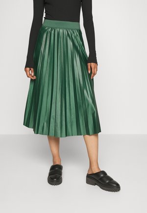 Femme portant un haut noir à manches longues, une jupe midi plissée verte et des chaussures plateformes noires sans lacets, debout sur un sol gris.
