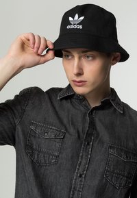 Cappello a secchiello in cotone nero con logo "adidas" bianco, indossato da una persona con una camicia di jeans grigio scuro con due tasche sul petto.