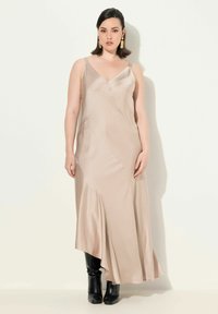 Beige Satin-Netz-Kleid mit V-Ausschnitt und verstellbaren Trägern, mit asymmetrischem Saum. Kombiniert mit glänzenden schwarzen kniehohen Stiefeln.