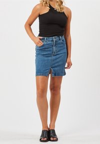 Femme portant un haut sans manches noir, une jupe en denim bleu avec une fente à l'avant et des chaussures à bout ouvert noires, debout contre un fond neutre.