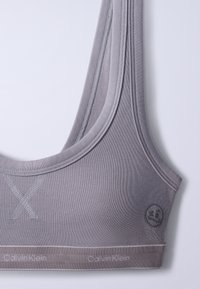 Κοντινή λήψη γκρι αθλητικού σουτιέν Calvin Klein με φαρδιές τιράντες, ριγέ ύφασμα και ραμμένη λεπτομέρεια X μπροστά.