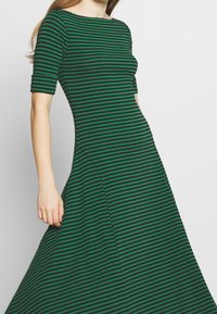 Kvinna i en grön och svart horisontellt randig klänning med ärmar som når till armbågen, stående mot en enkel ljus bakgrund.
