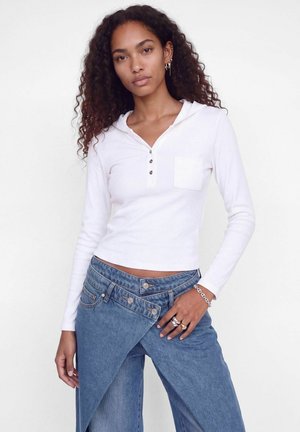 Un mannequin pose sur fond blanc, vêtu d’un haut blanc à manches longues et à capuche, ainsi que d’un jean bleu à devant portefeuille.