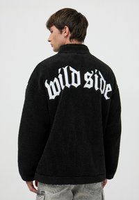 Czarna polarowa kurtka o puszystej fakturze, z napisem "dzika strona" dużymi białymi literami z tyłu. Stylizowana z wysokim kołnierzem.
