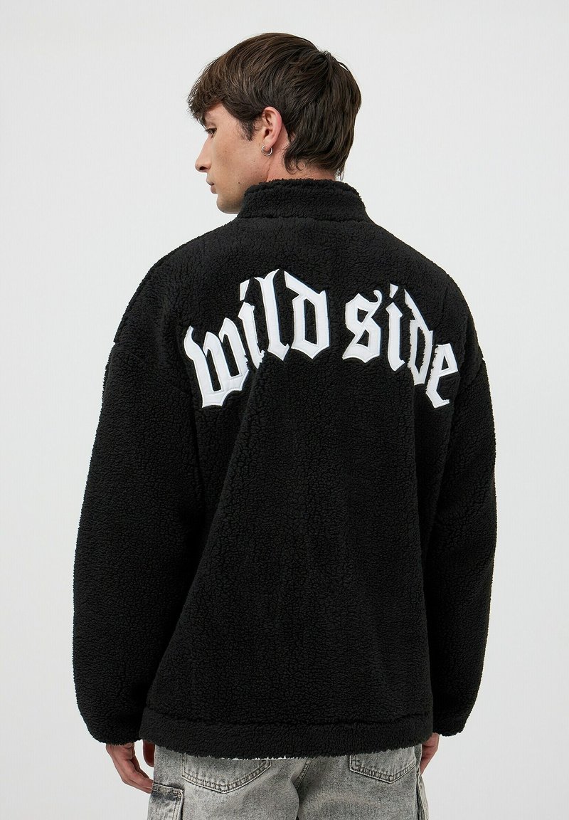Czarna polarowa kurtka o puszystej fakturze, z napisem "dzika strona" dużymi białymi literami z tyłu. Stylizowana z wysokim kołnierzem.