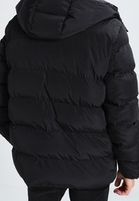 Giubbotto a vento nero con cappuccio, caratterizzato da un design trapuntato e canali orizzontali. Realizzato in materiale sintetico, offre una texture liscia.