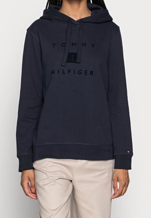 Osoba ubrana w granatową bluzę z kapturem Tommy Hilfiger ze sznurkami i beżowe spodnie, stojąca na jasnym tle.