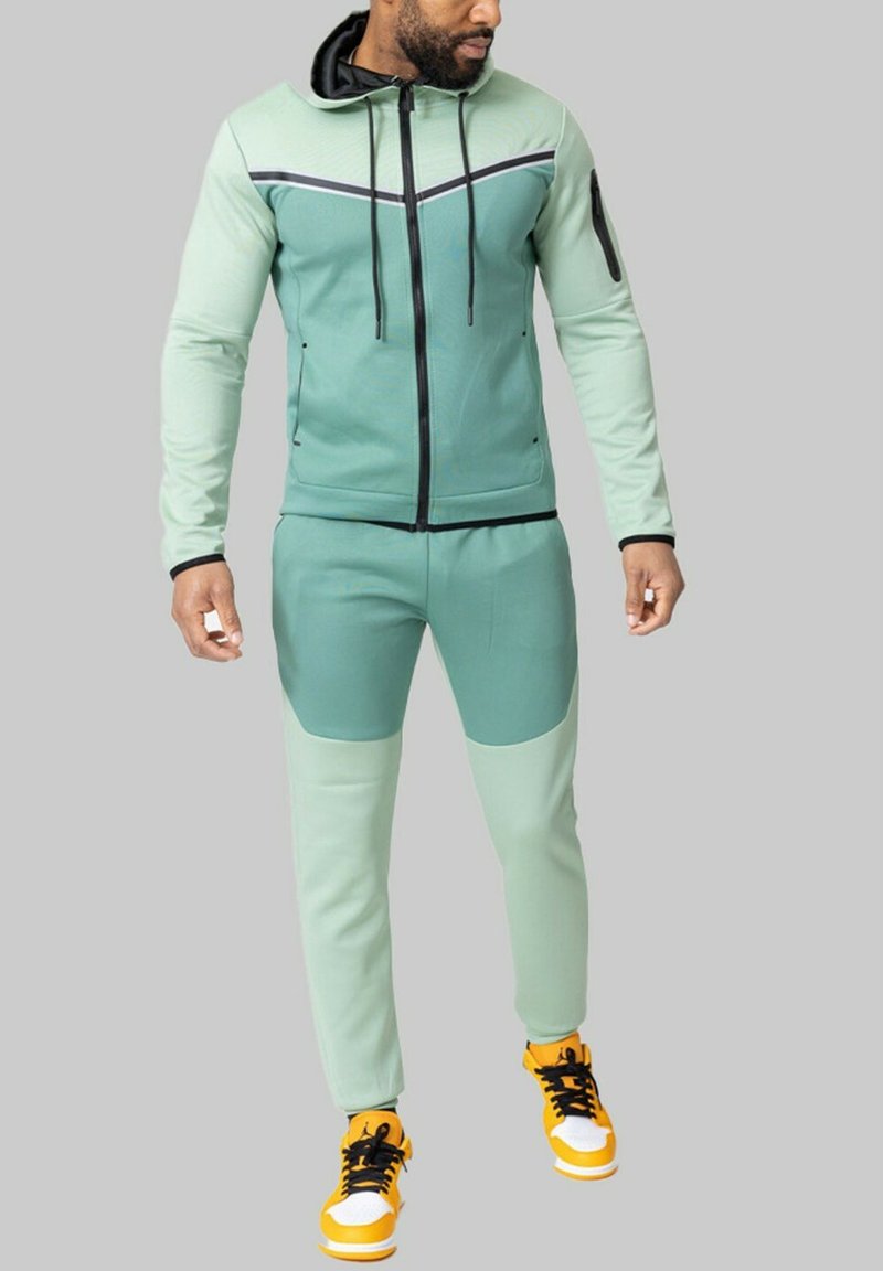 Ensemble de sport vert avec capuche, présentant des accents noirs contrastés et des poches zippées. Associé à des baskets jaunes vives.