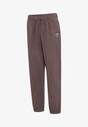 Barn iført brun fleece træningsdragt, sort hue og sorte sneakers, stående med hænderne bag ryggen på ensfarvet hvid baggrund.