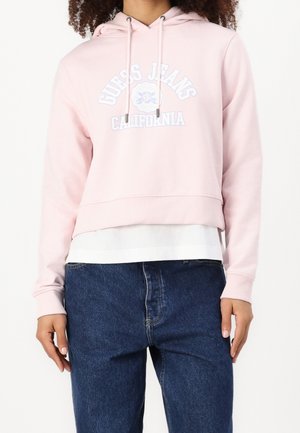 Osoba nosząca jasnoróżową bluzę z kapturem "Guess Jeans California" na białej koszuli i luźnych ciemnoniebieskich dżinsach, pokazana od szyi do bioder.