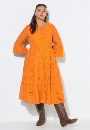 Woman wearing a bright orange long-sleeve dress with eyelet patterns and brown embroidered cowboy boots, standing against a white background.